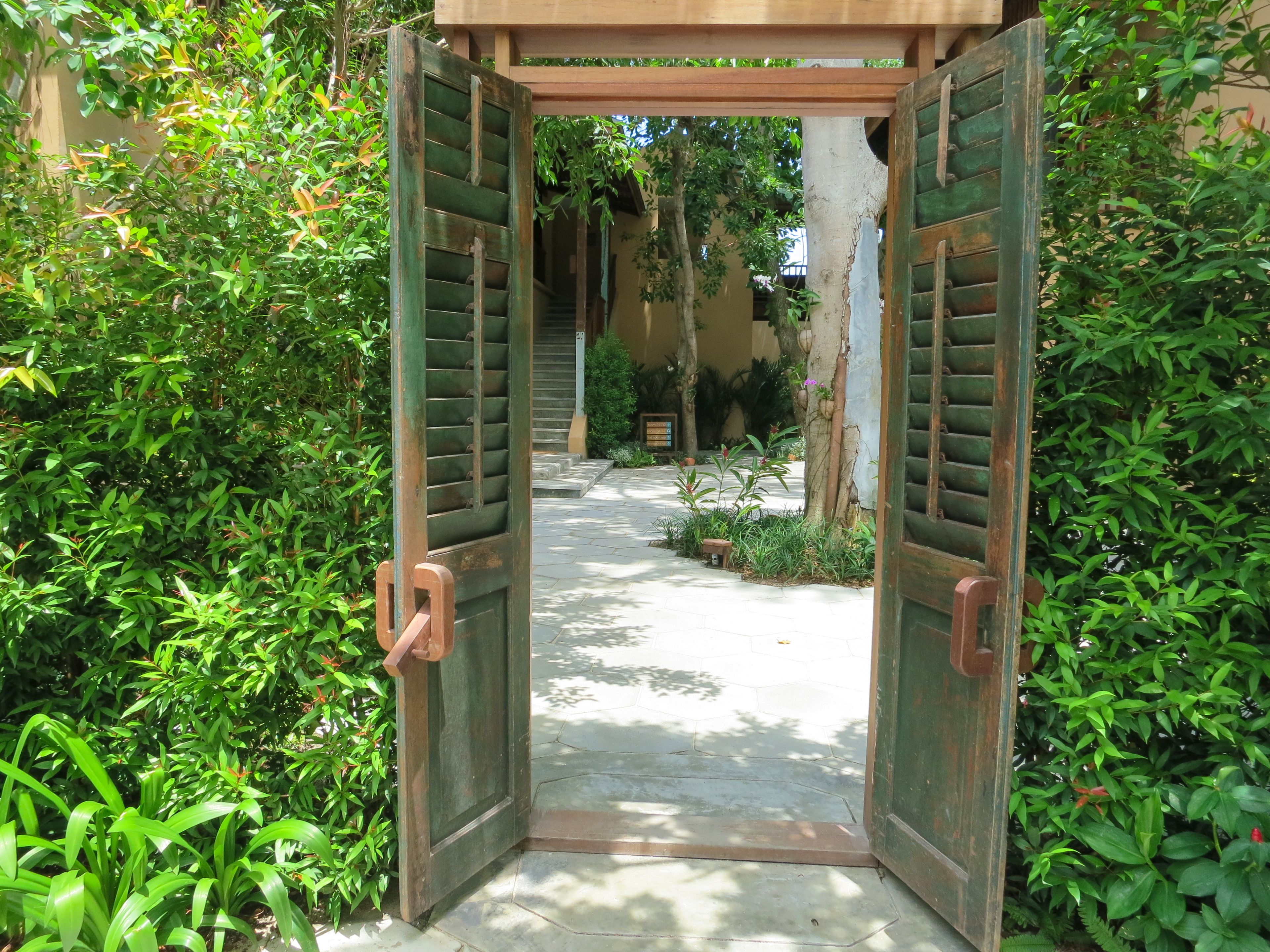 An open wooden gate leading to a paved courtyard surrounded by lush greenery.