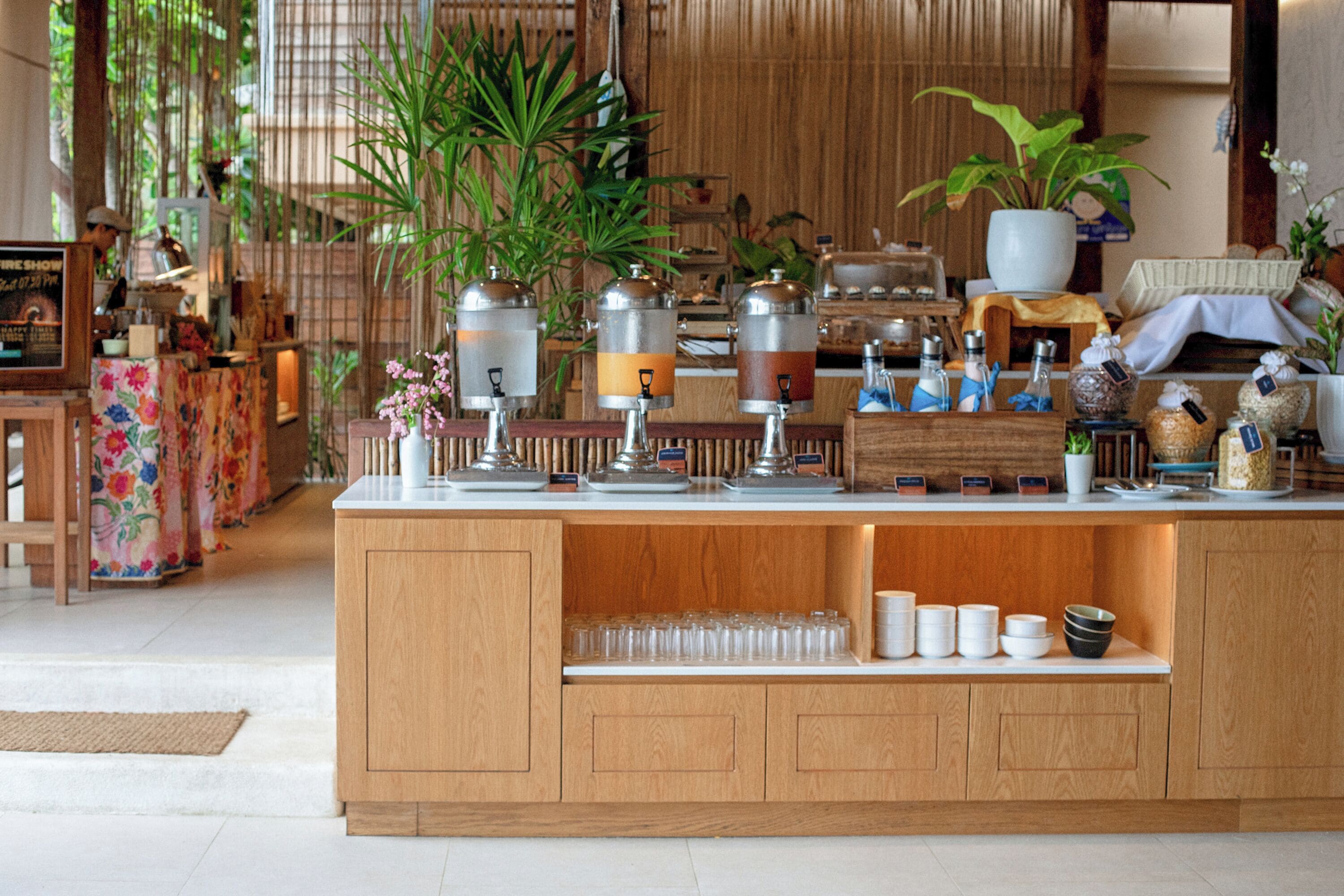 A wooden bar area with a variety of drink dispensers, a potted plant, and a display of glassware.