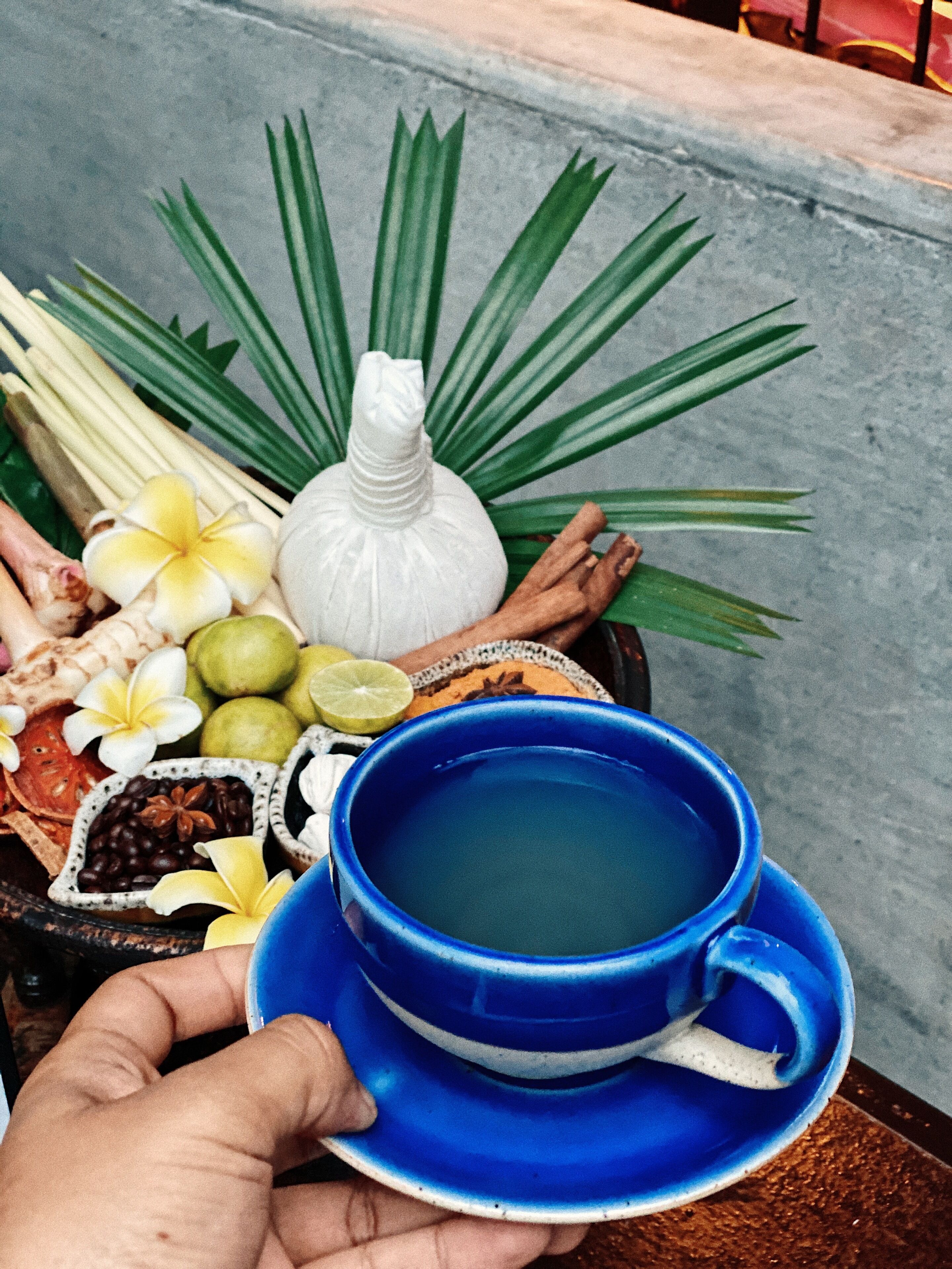 A hand holding a blue cup with a saucer, filled with a green liquid, surrounded by various items including a white bottle, green leaves, and small bowls containing different ingredients.
