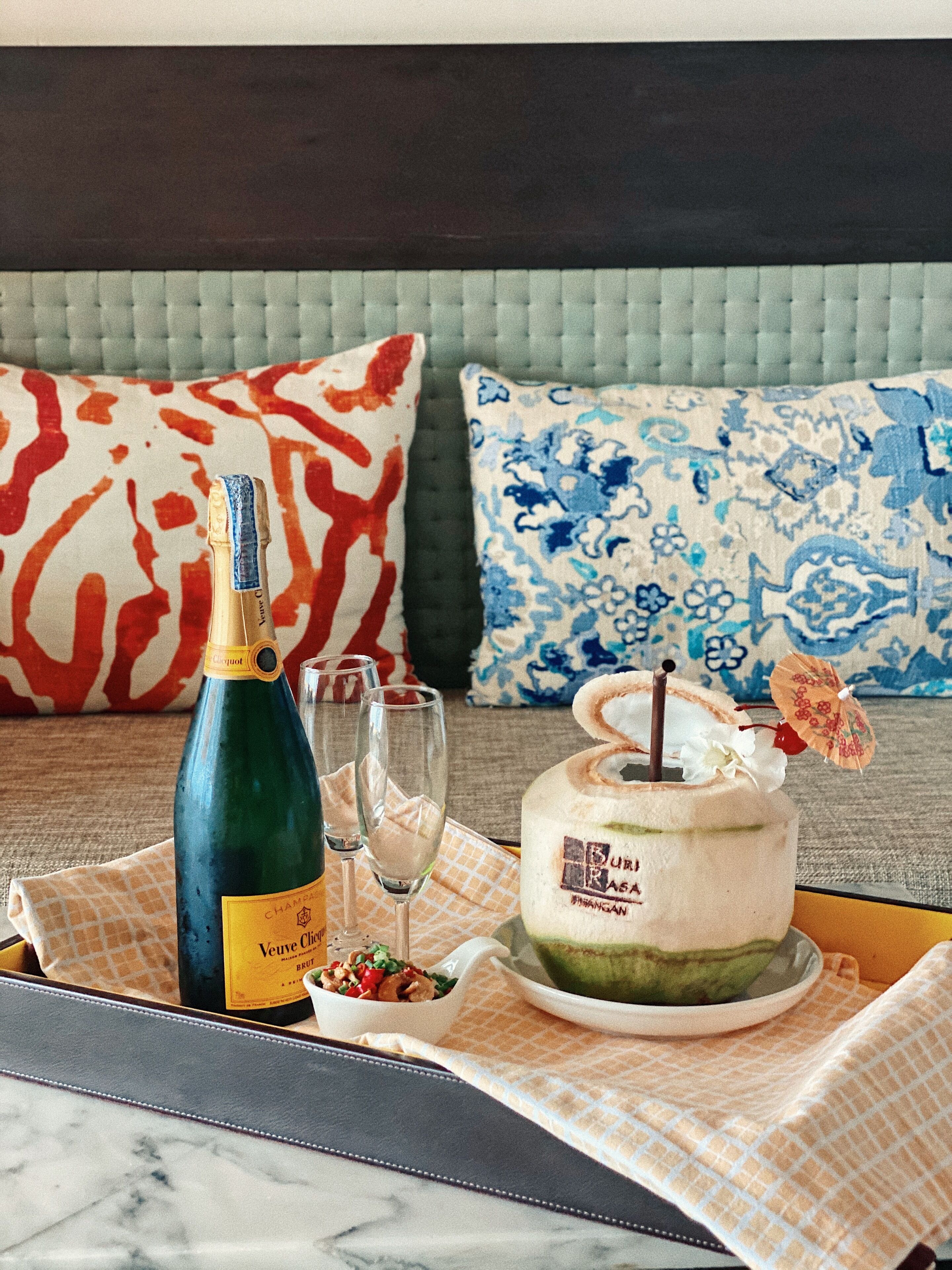 A dining table with a bottle of Veuve Clicquot champagne, two glasses, a cake with a cherry on top, and a bowl of fruit.