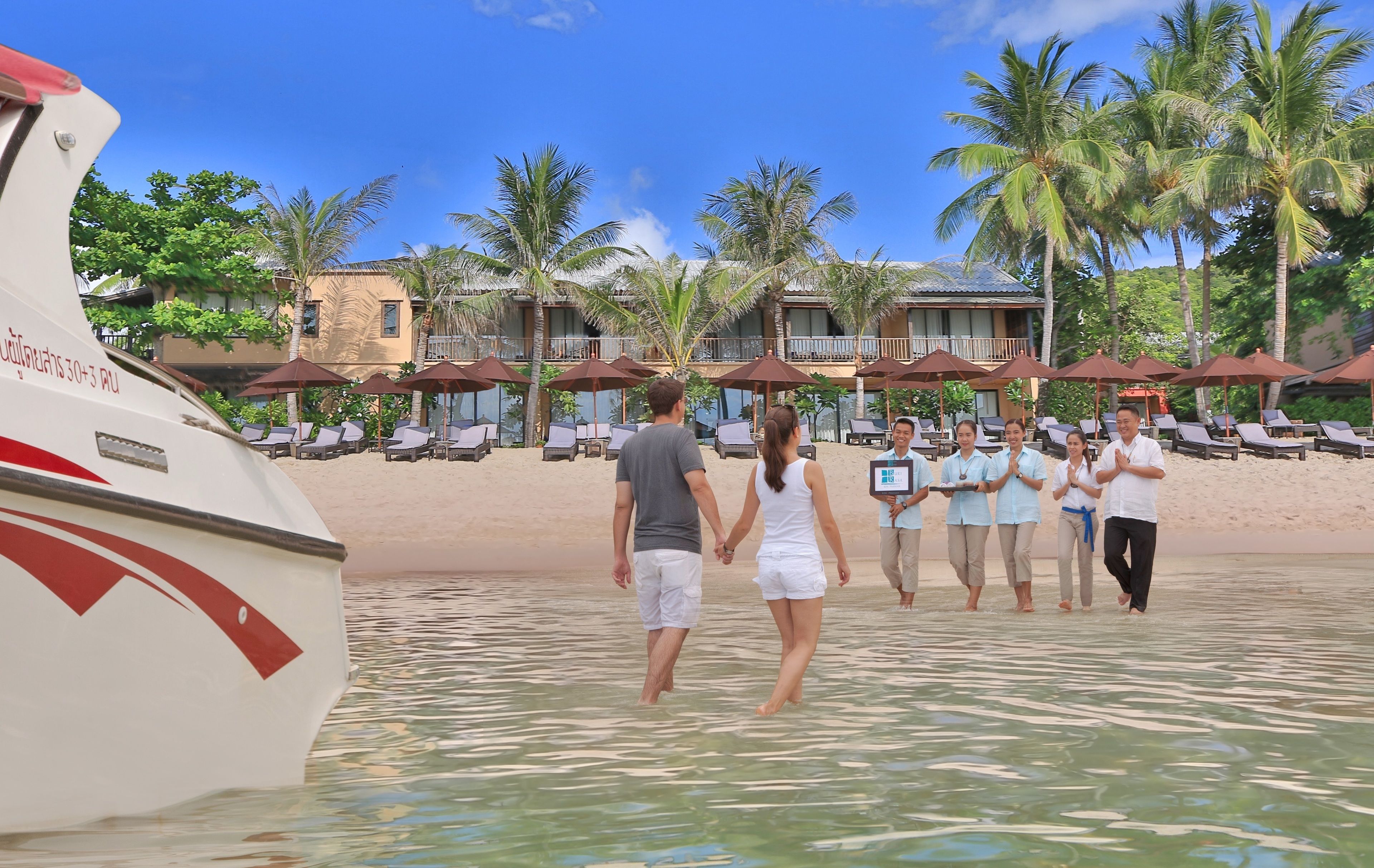 A beachfront hotel with a poolside area, palm trees, and a boat docked nearby.
