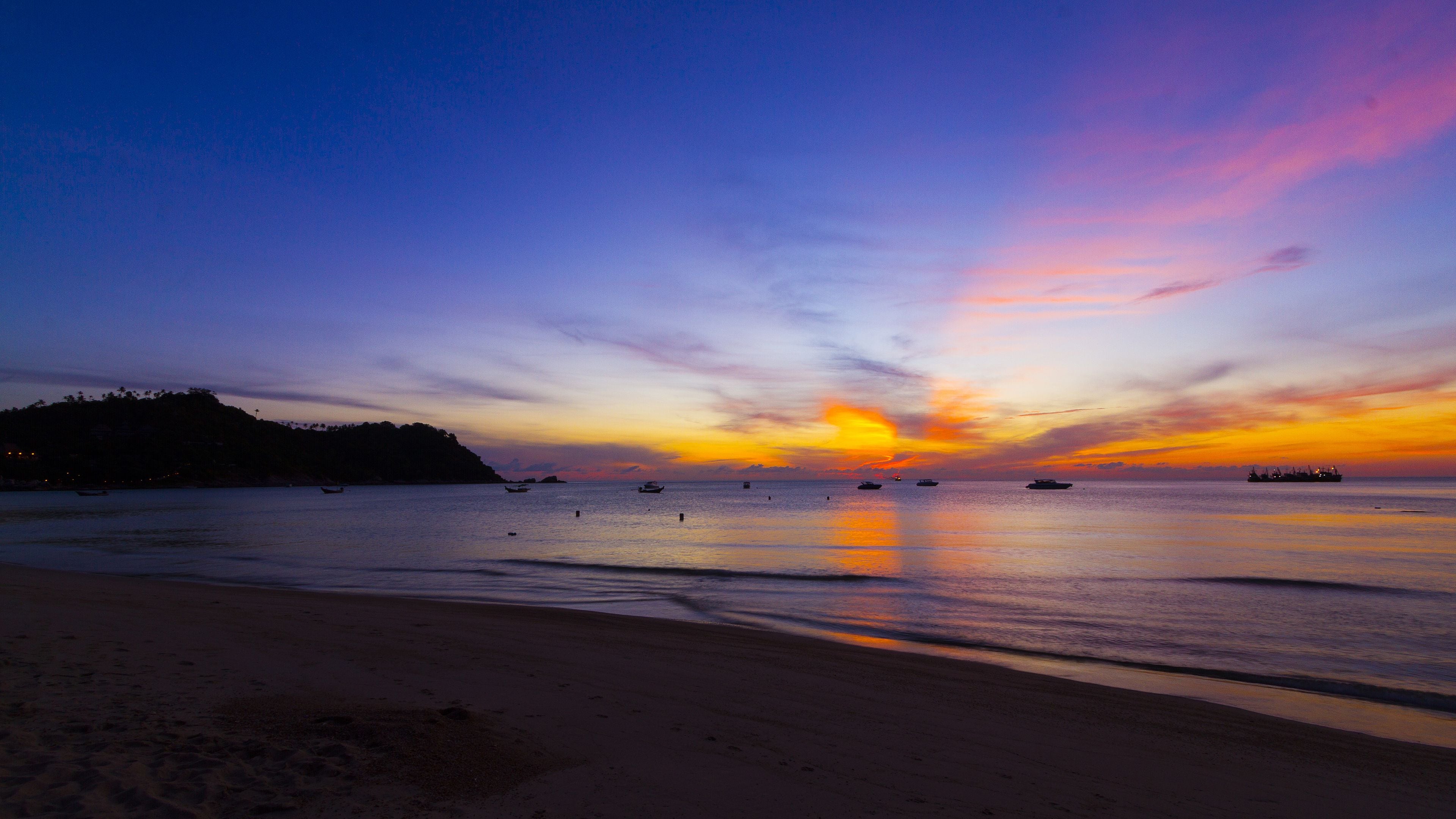 A beach at sunset with boats on the water and a distant landmass.