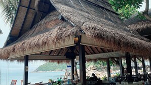 A beachside bar with a thatched roof, wooden chairs, and a view of the sea. - Buri Rasa Village Phangan (Ko Pha-ngan)