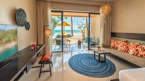 A hotel room with a large TV, a sofa with colorful cushions, a dining table, and a view of the beach through large windows. - Buri Rasa Village Phangan (Ko Pha-ngan)