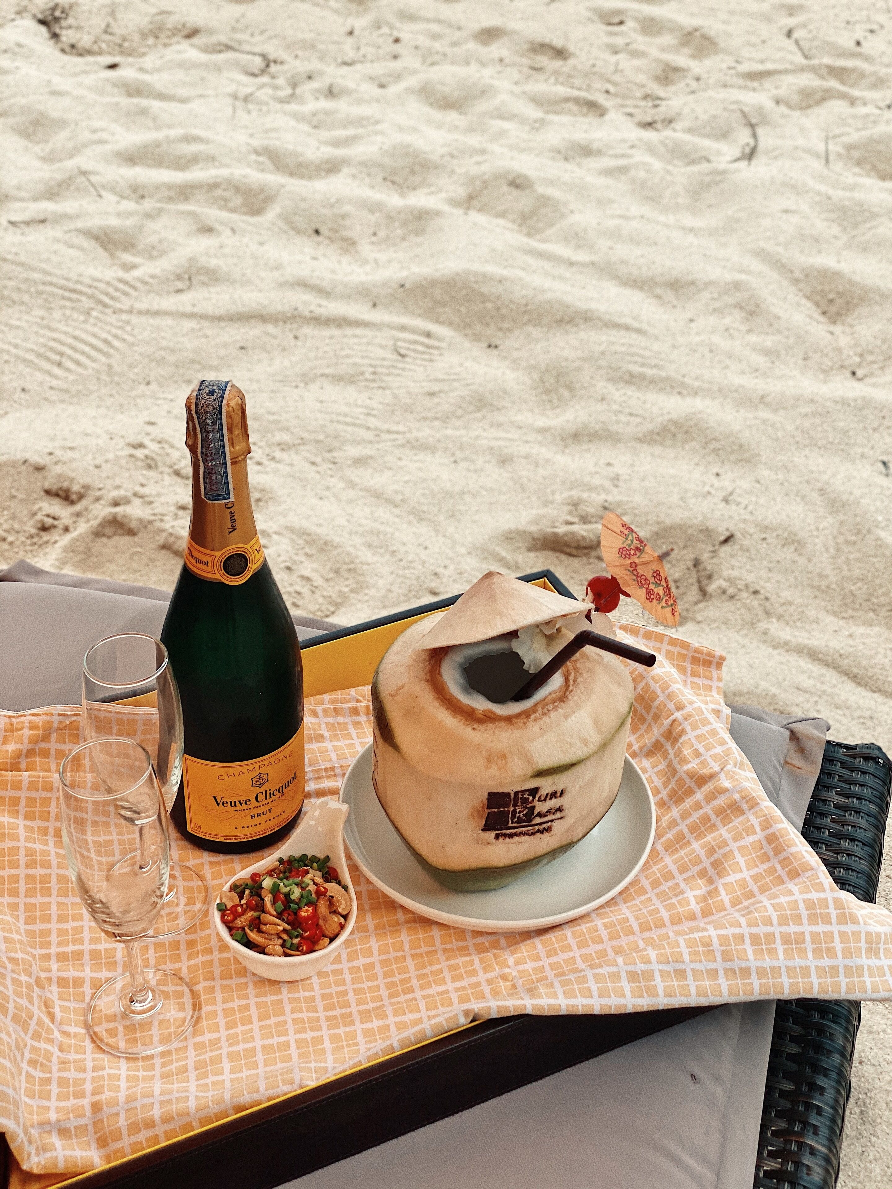 A bottle of Veuve Clicquot champagne, two glasses, a coconut dessert, and a small bowl of nuts on a checkered tablecloth on a beach.