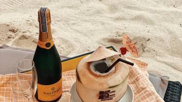 A bottle of Veuve Clicquot champagne, two glasses, a coconut dessert, and a small bowl of nuts on a checkered tablecloth on a beach.
