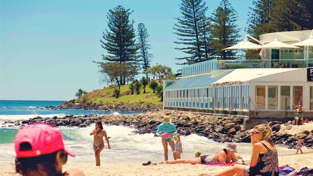 A beach with people sunbathing and walking. There are trees and a lifeguard tower in the background. - Key Largo Apartments (Burleigh Heads)