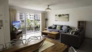A living room with a wooden dining table, grey sofa, and a TV on a stand. - Key Largo Apartments (Burleigh Heads)