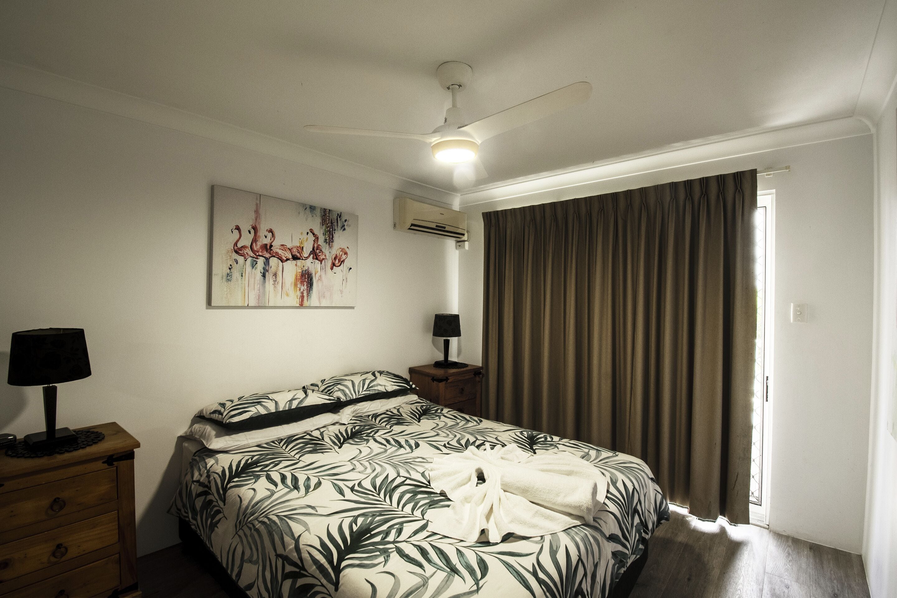 A bedroom with a bed, a ceiling fan, an air conditioning unit, a nightstand with a lamp, and a window with curtains.