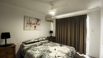 A bedroom with a bed, a ceiling fan, an air conditioning unit, a nightstand with a lamp, and a window with curtains.