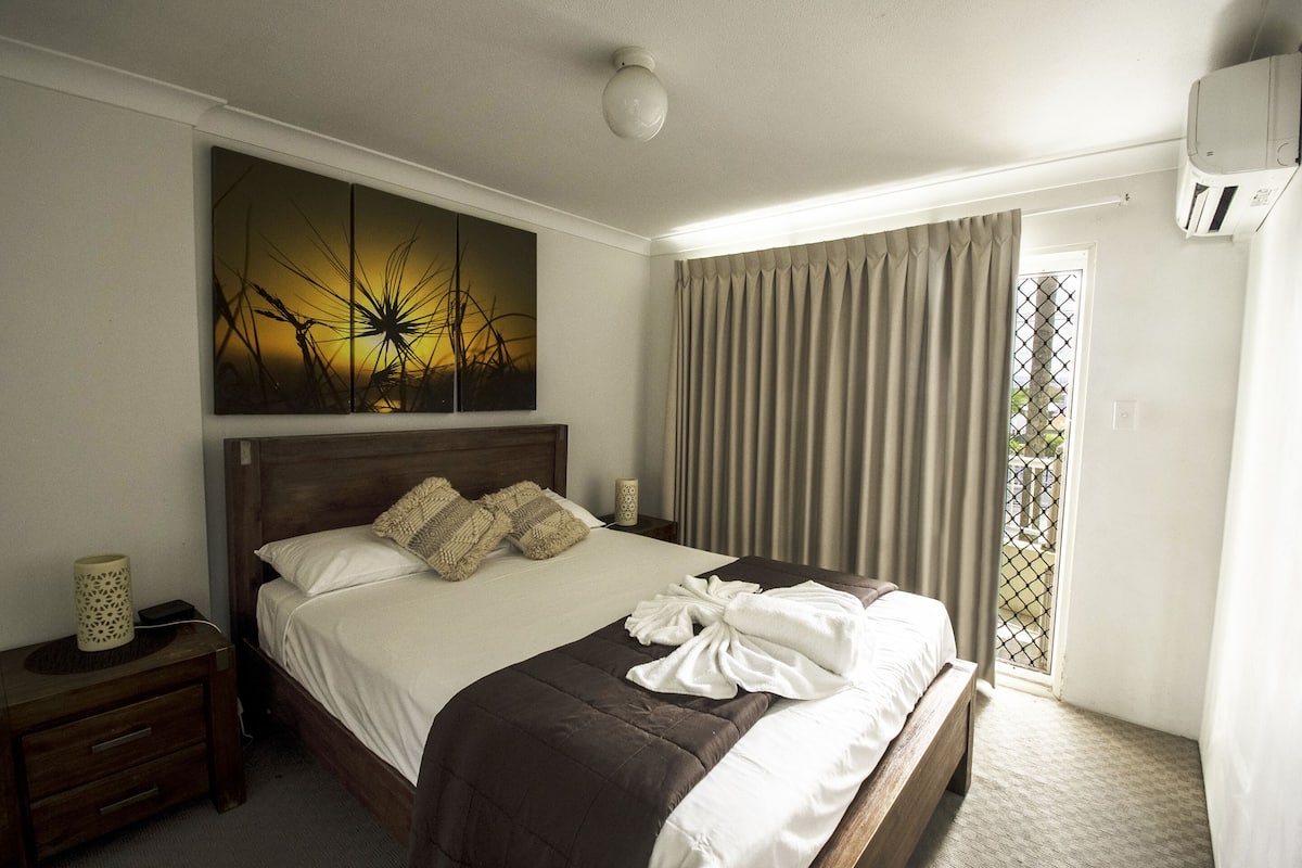 A hotel room with a large bed, a nightstand, a wall-mounted air conditioner, and a window with curtains.