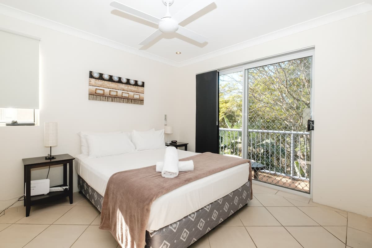 Une chambre d’hôtel avec un grand lit, un ventilateur de plafond, une fenêtre donnant sur la verdure et une table de chevet avec une lampe.