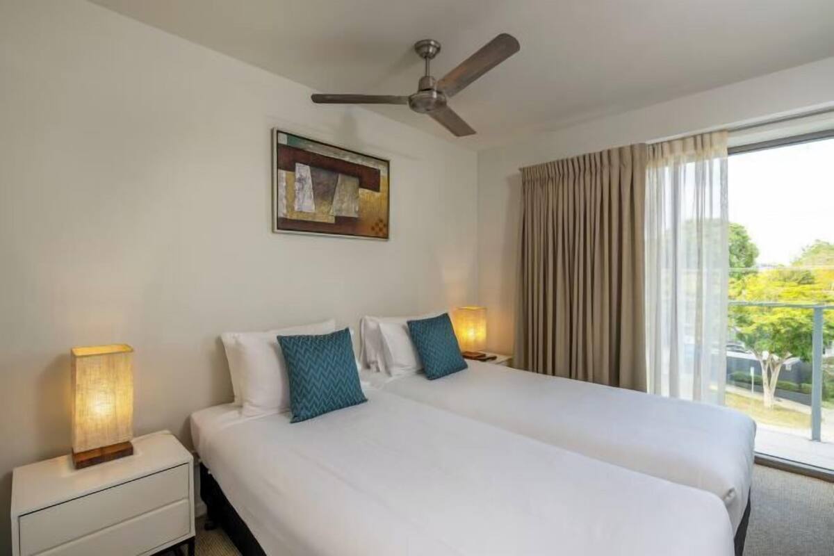 A hotel room with two beds, a ceiling fan, a window with curtains, and a wall-mounted artwork.
