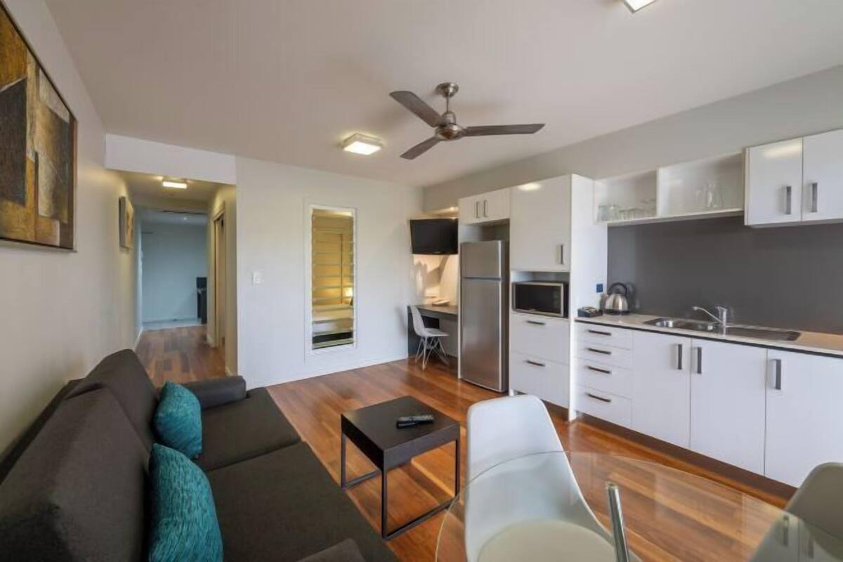 A modern living room with a sofa, a small table, and a TV. There is a kitchen with white cabinets and a refrigerator.