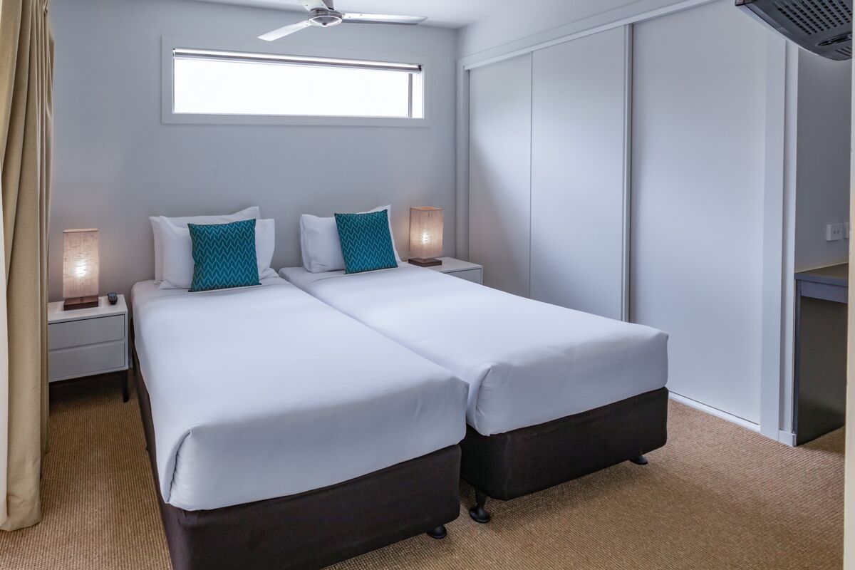 Ein Doppelbett mit weißen und türkisen Kissen, ein Deckenventilator, ein Nachttisch mit Lampe und ein Fenster mit Jalousien.