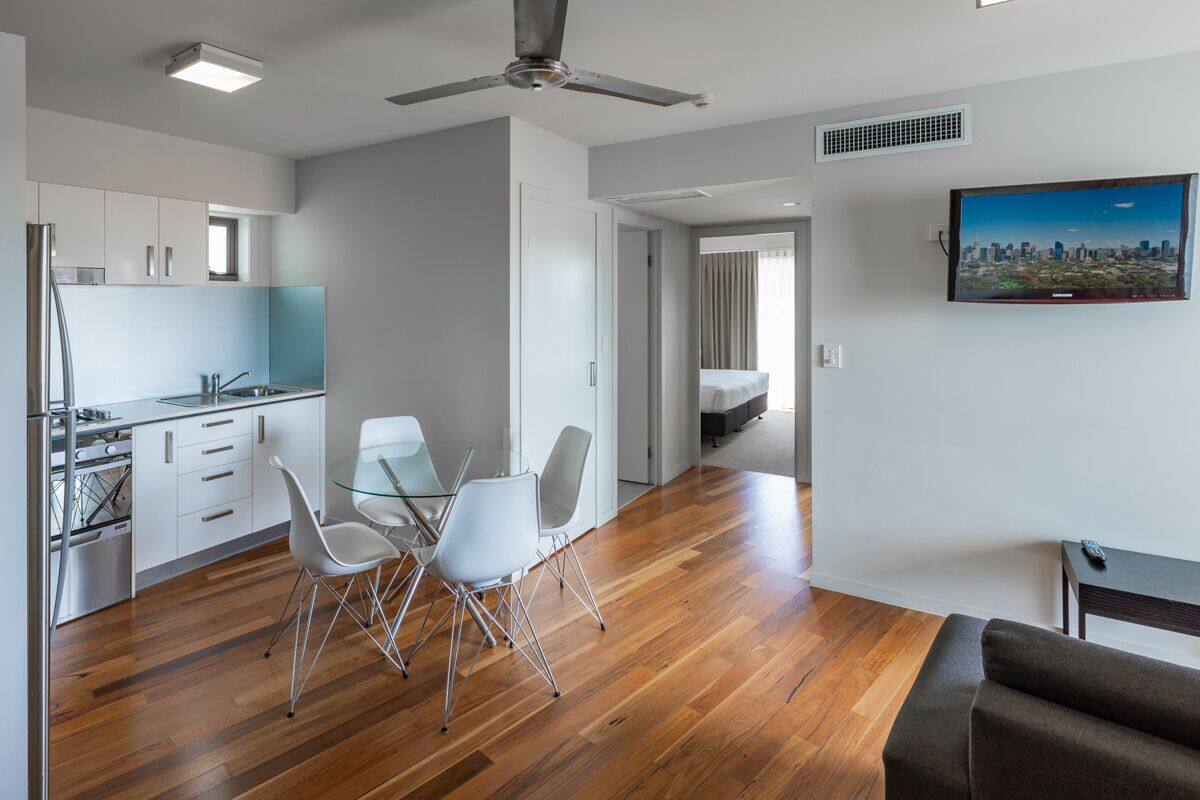 A modern hotel room with a wooden floor, a dining area with white chairs, a bed, a TV, and a ceiling fan.