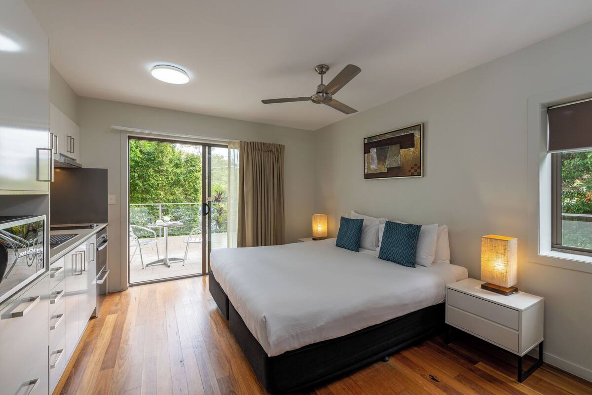 A modern bedroom with a large bed, a ceiling fan, a bedside lamp, and a view of the outdoors through a sliding door.