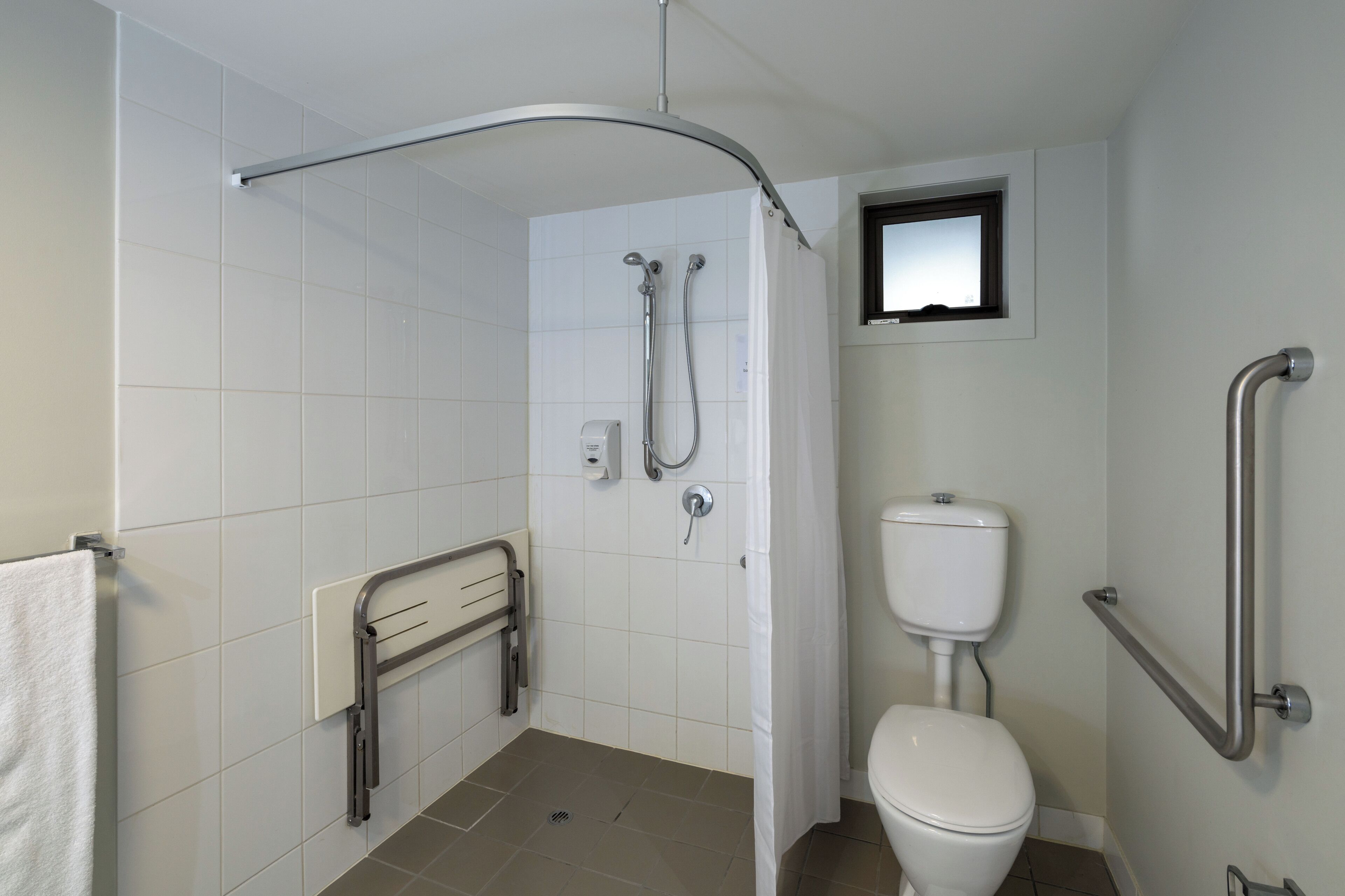 A bathroom with white tiles, a white toilet, a shower with a curved glass door, and a small window above the toilet.