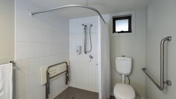 A bathroom with white tiles, a white toilet, a shower with a curved glass door, and a small window above the toilet.