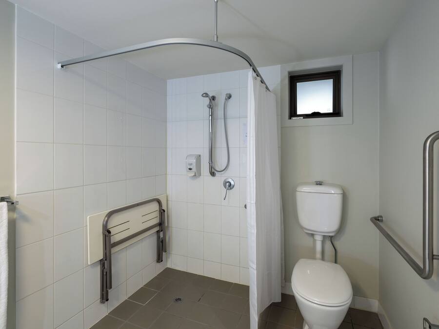 A bathroom with white tiles, a white toilet, a shower with a curved glass door, and a small window above the toilet.
