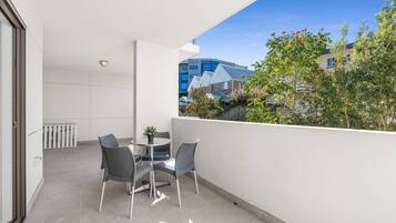 A balcony with a table and chairs, a potted plant, and a view of neighboring buildings and trees.