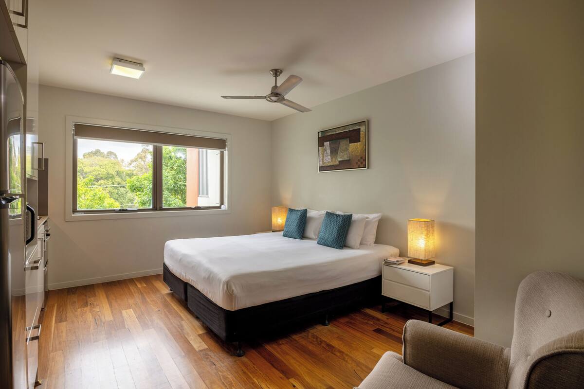 A bedroom with a large bed, a ceiling fan, a bedside lamp, and a window with a view of greenery.
