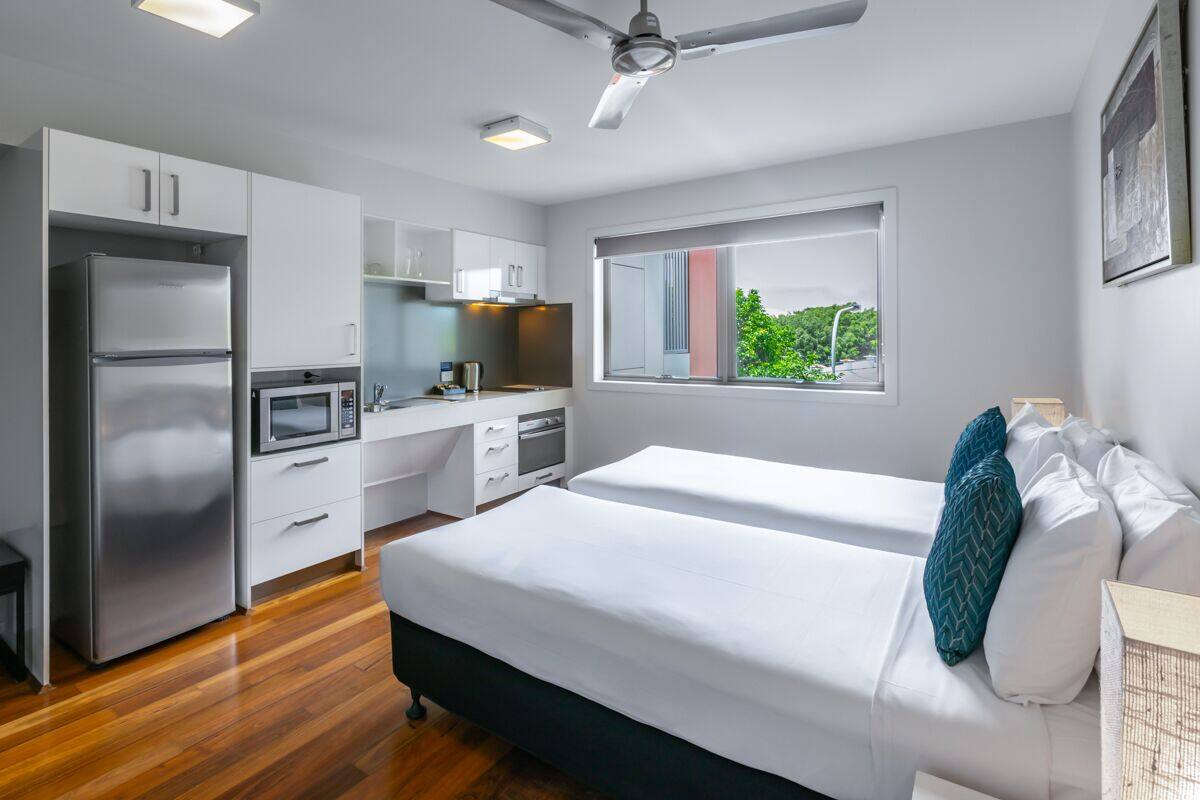 A modern hotel room with a kitchenette, a bed with white bedding, a window with a view, and a ceiling fan.