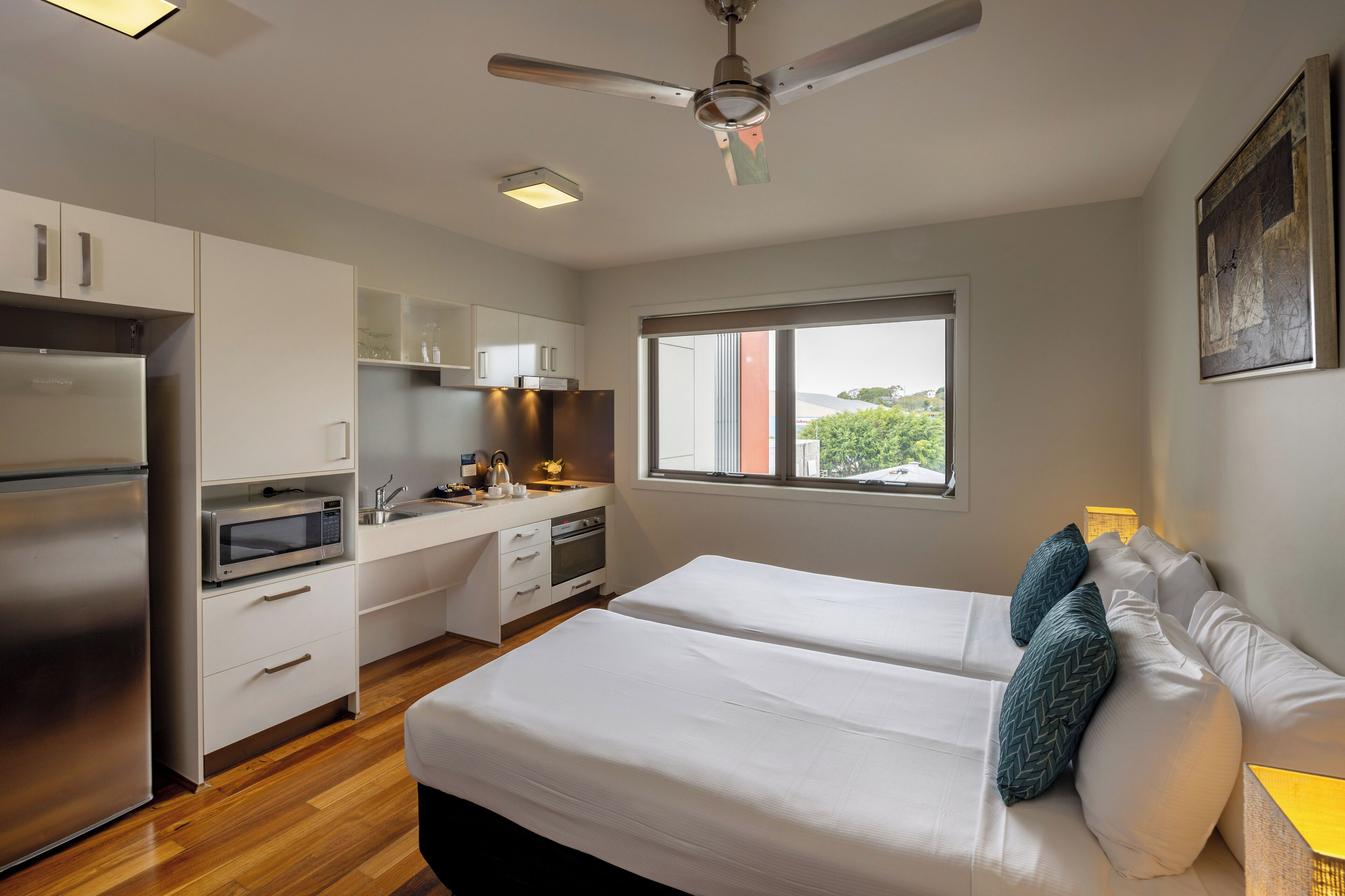 Una habitación de hotel moderna con kitchenette, una cama con ropa blanca y almohadones decorativos, un ventilador de techo y una ventana con vistas a zonas verdes.