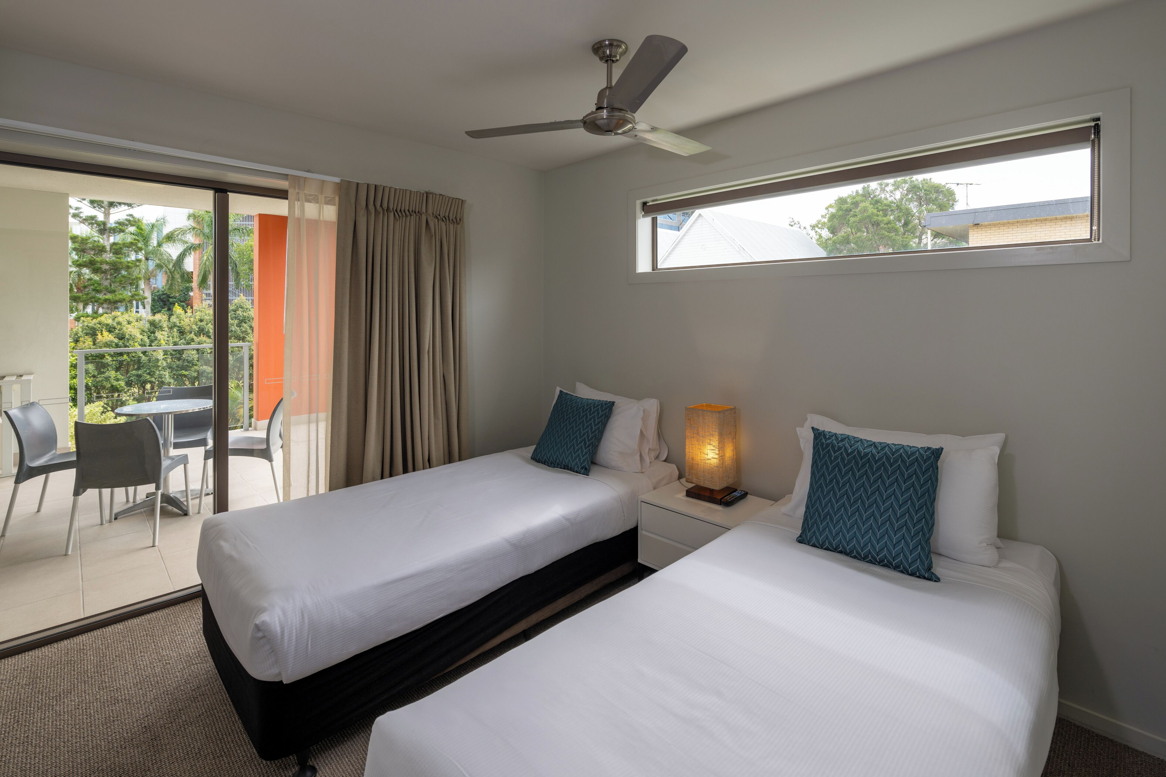 Habitación de hotel con dos camas, ventilador de techo, ventana y balcón con mobiliario exterior.