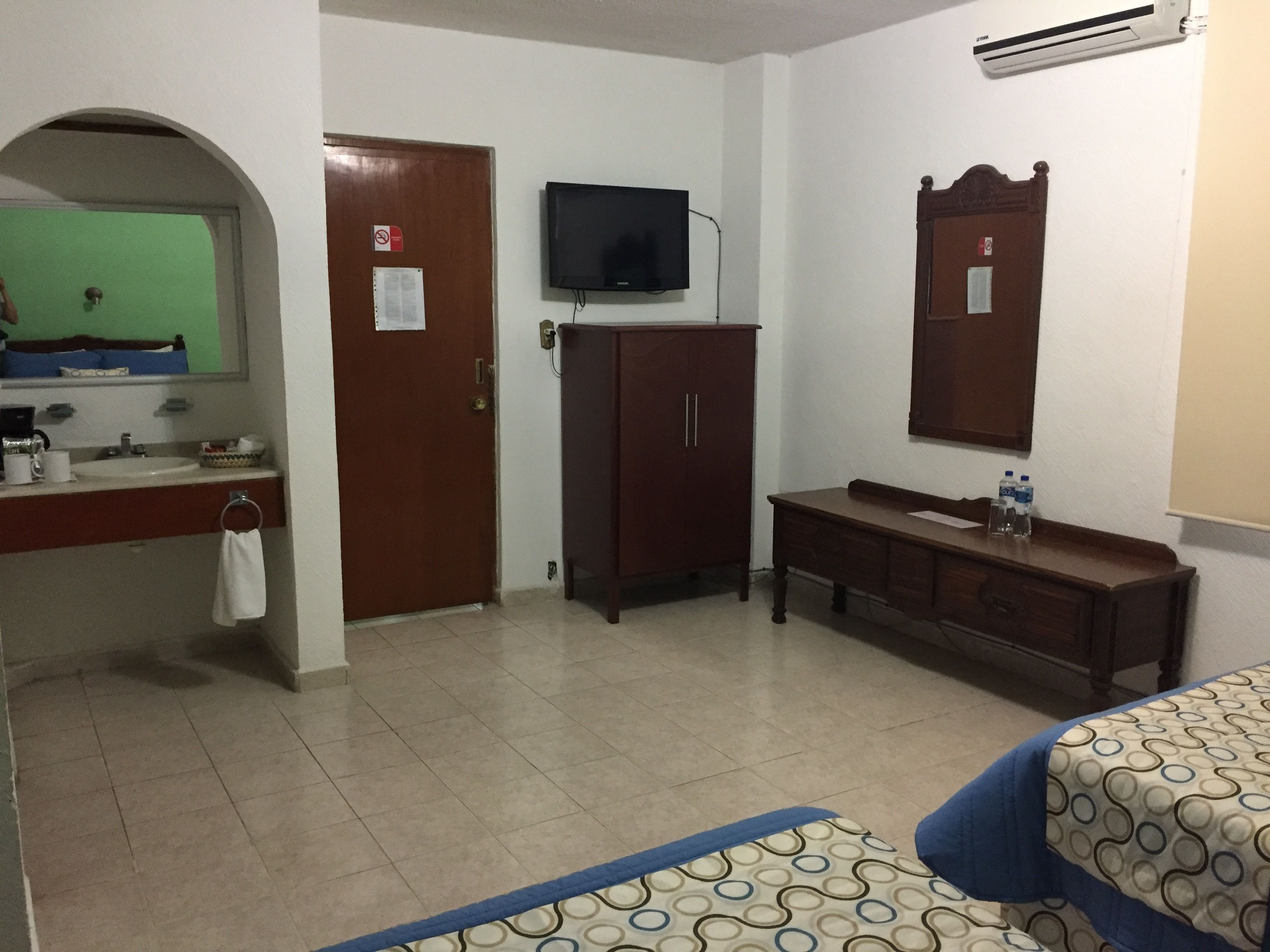 Habitación de hotel con cama, cómoda, televisor, espejo, puerta y baño con lavabo y espejo.