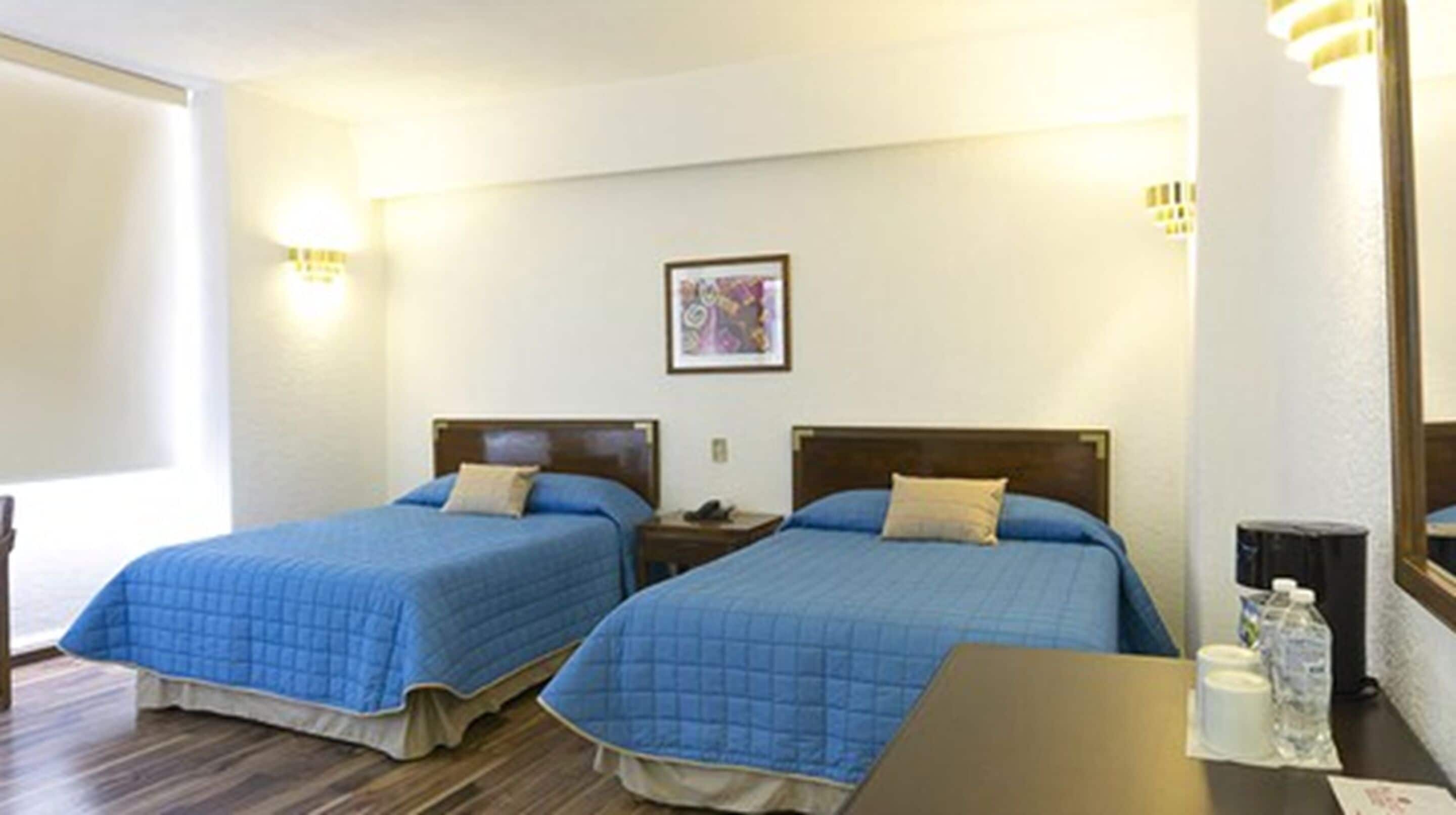 Una camera d'albergo con due letti, un tavolino, una sedia, un comodino, un quadro appeso al muro e una finestra.