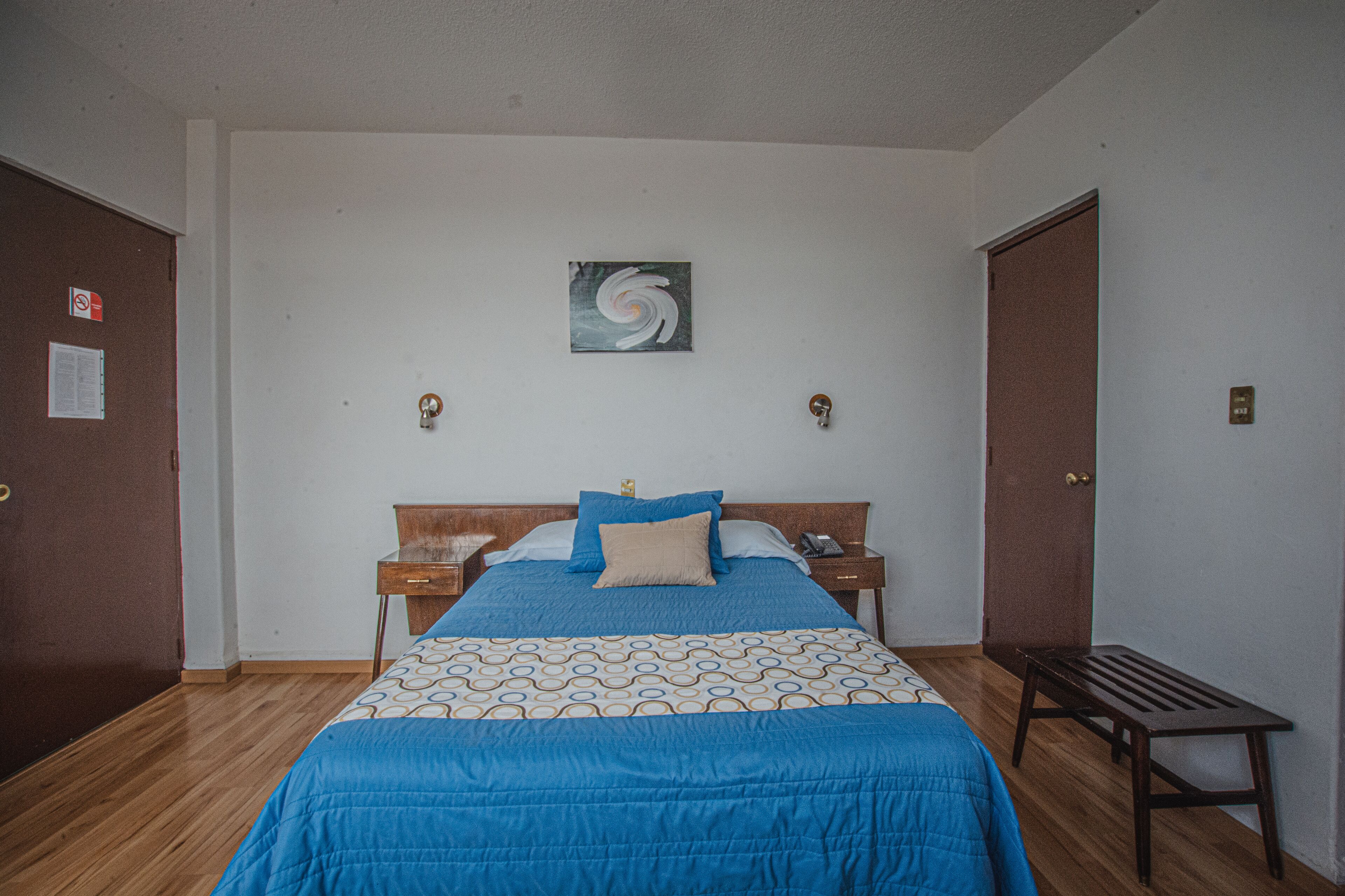 Un dormitorio con una cama, una mesita de noche, un banco, una puerta y una obra de arte colgada en la pared.