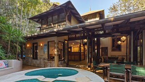 A pool area with a curved design, a dining area with green chairs, and a building with a balcony and large windows. - The Garden Burees (Byron Bay)