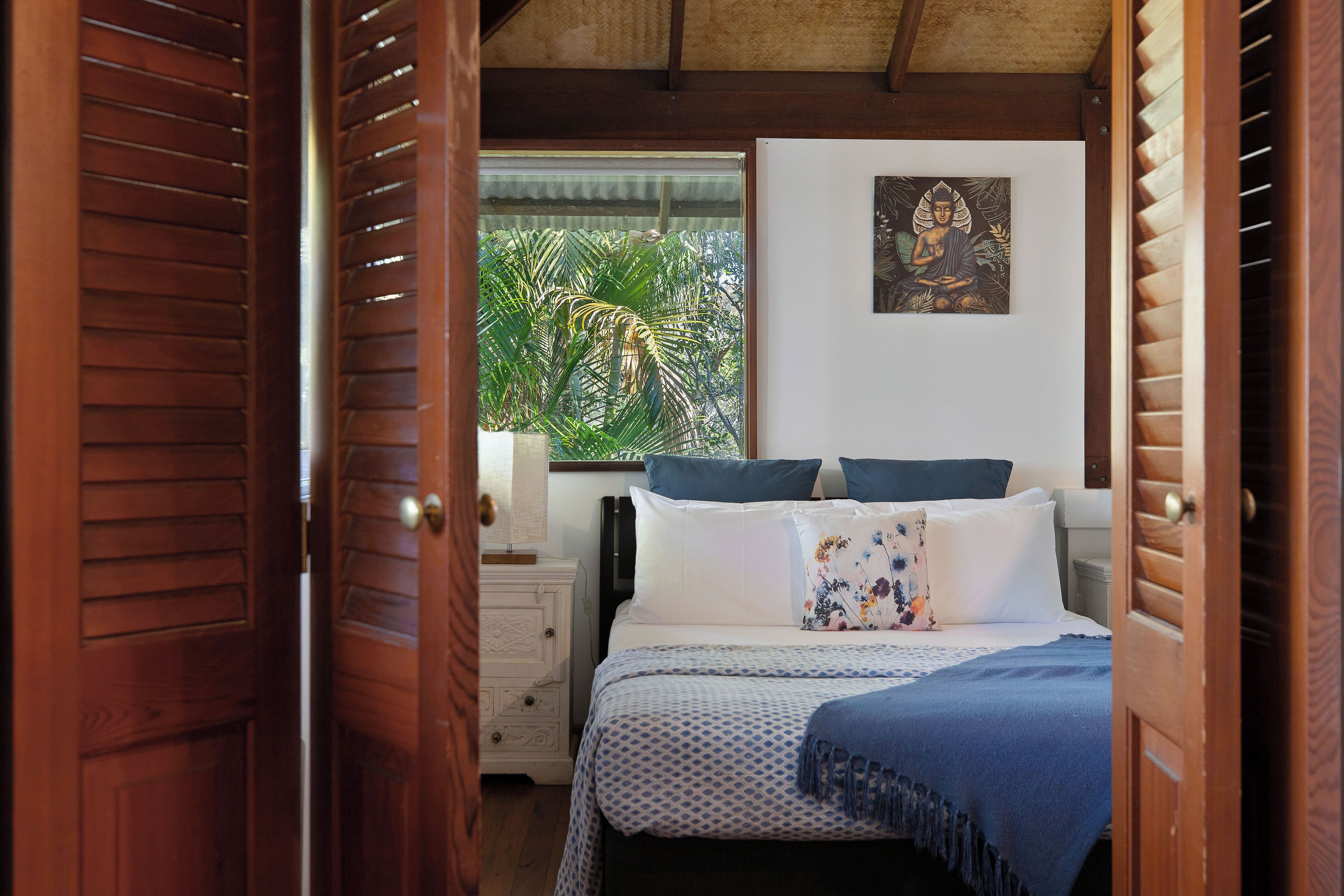 A bedroom with a bed, a nightstand, a window with a view of greenery, and a wall-mounted artwork.