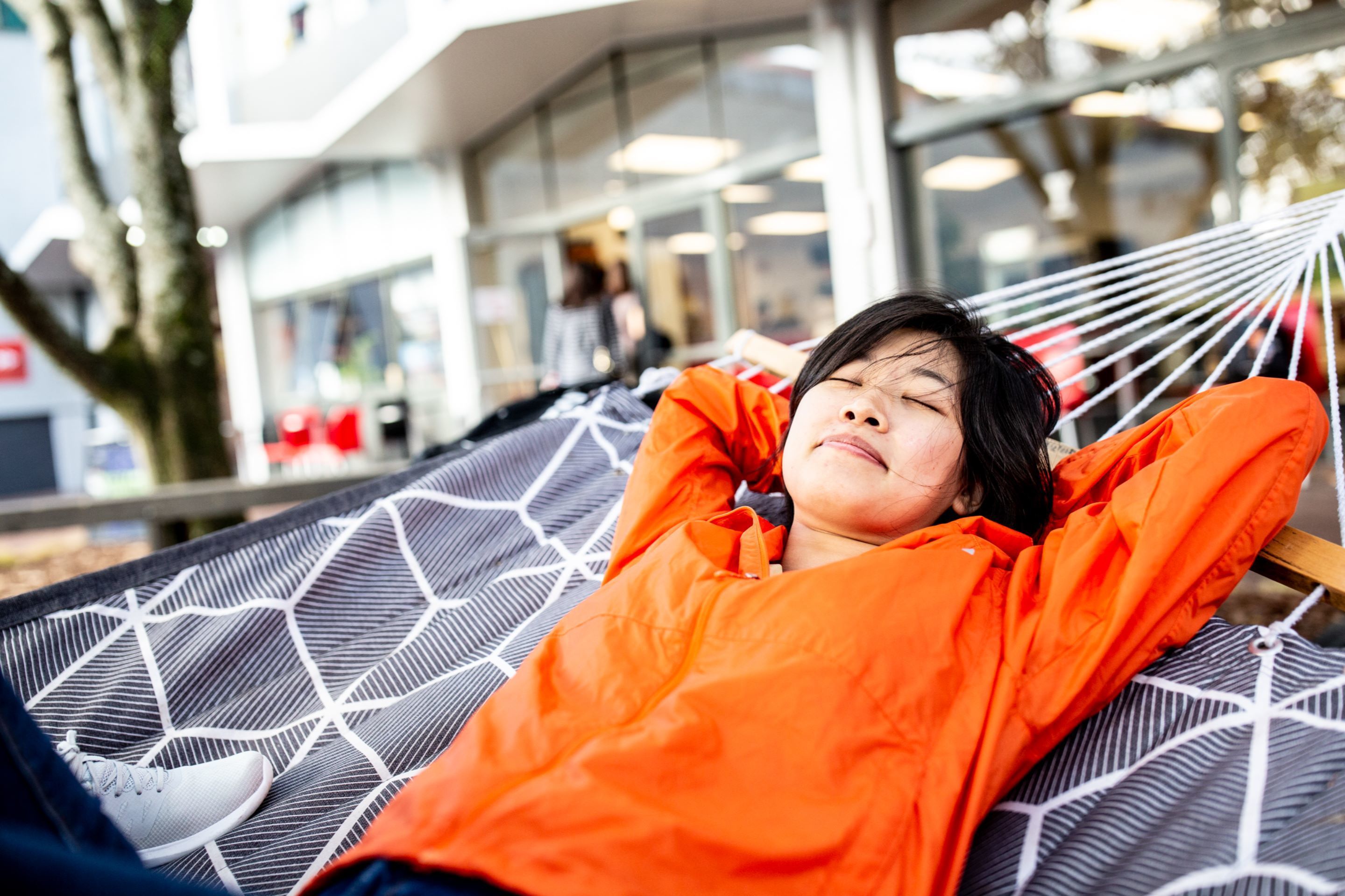 A person lying in a hammock with a geometric patterned fabric, outdoors with a modern building in the background.