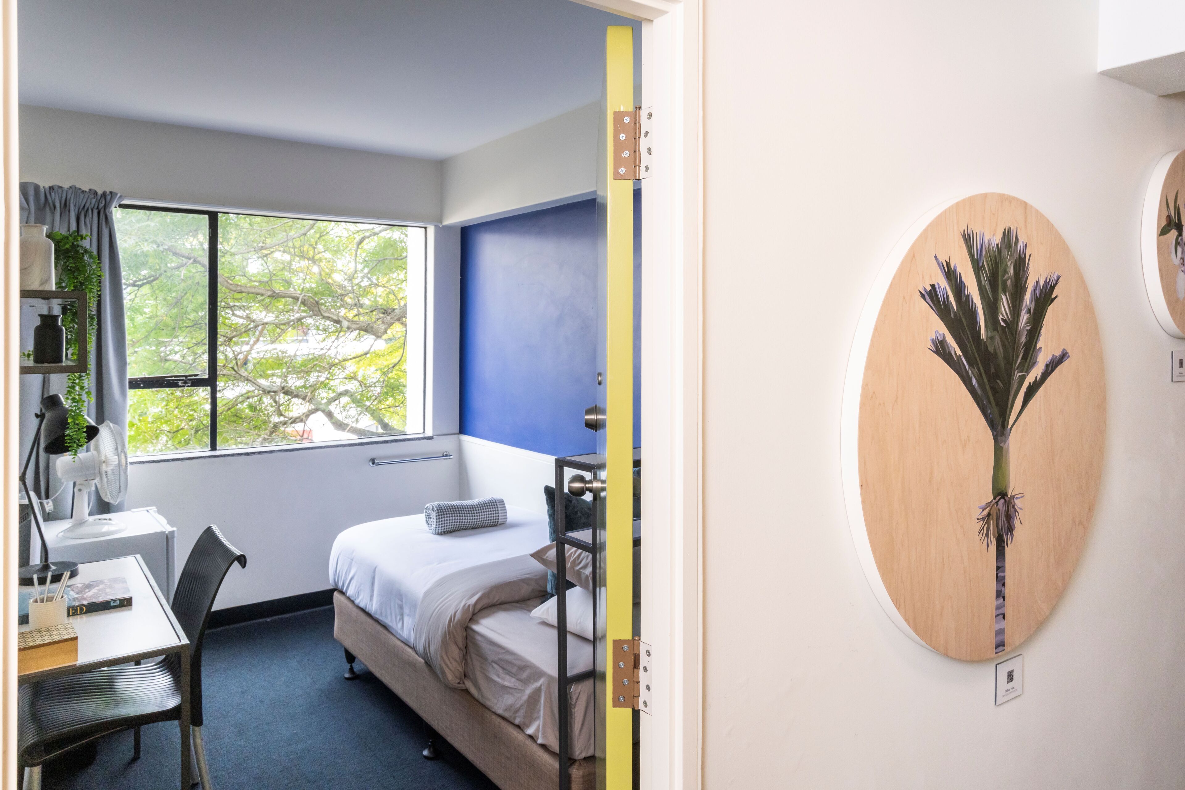 A room with a bed, a desk, a chair, a window with curtains, and a wall-mounted artwork.