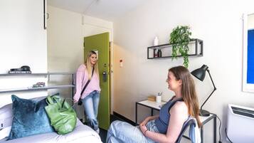 A hotel room with a bed, a desk with a computer, a green door, and a shelf with plants.
