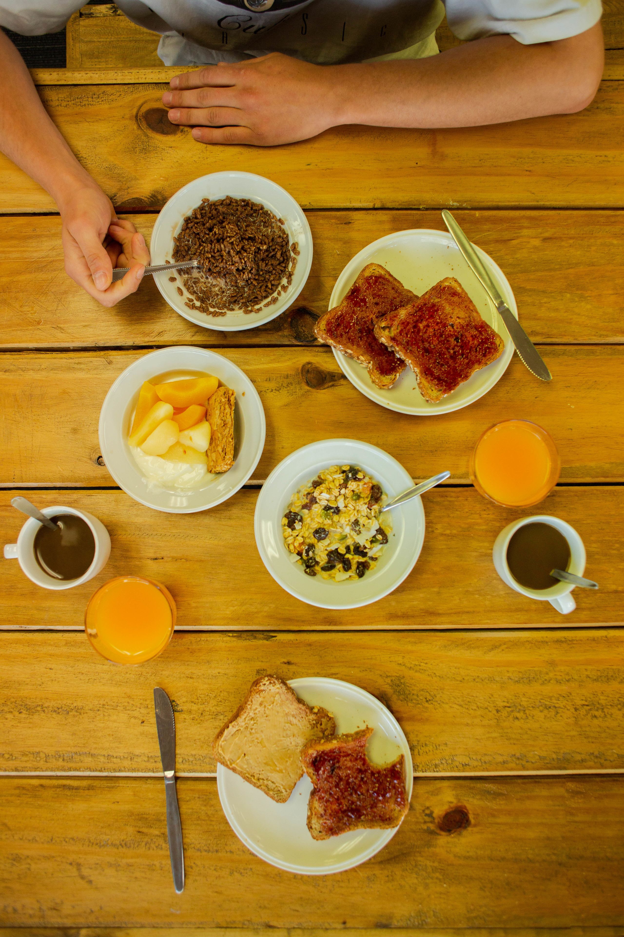 A wooden table with breakfast items including toast, fruit, cereal, and coffee.