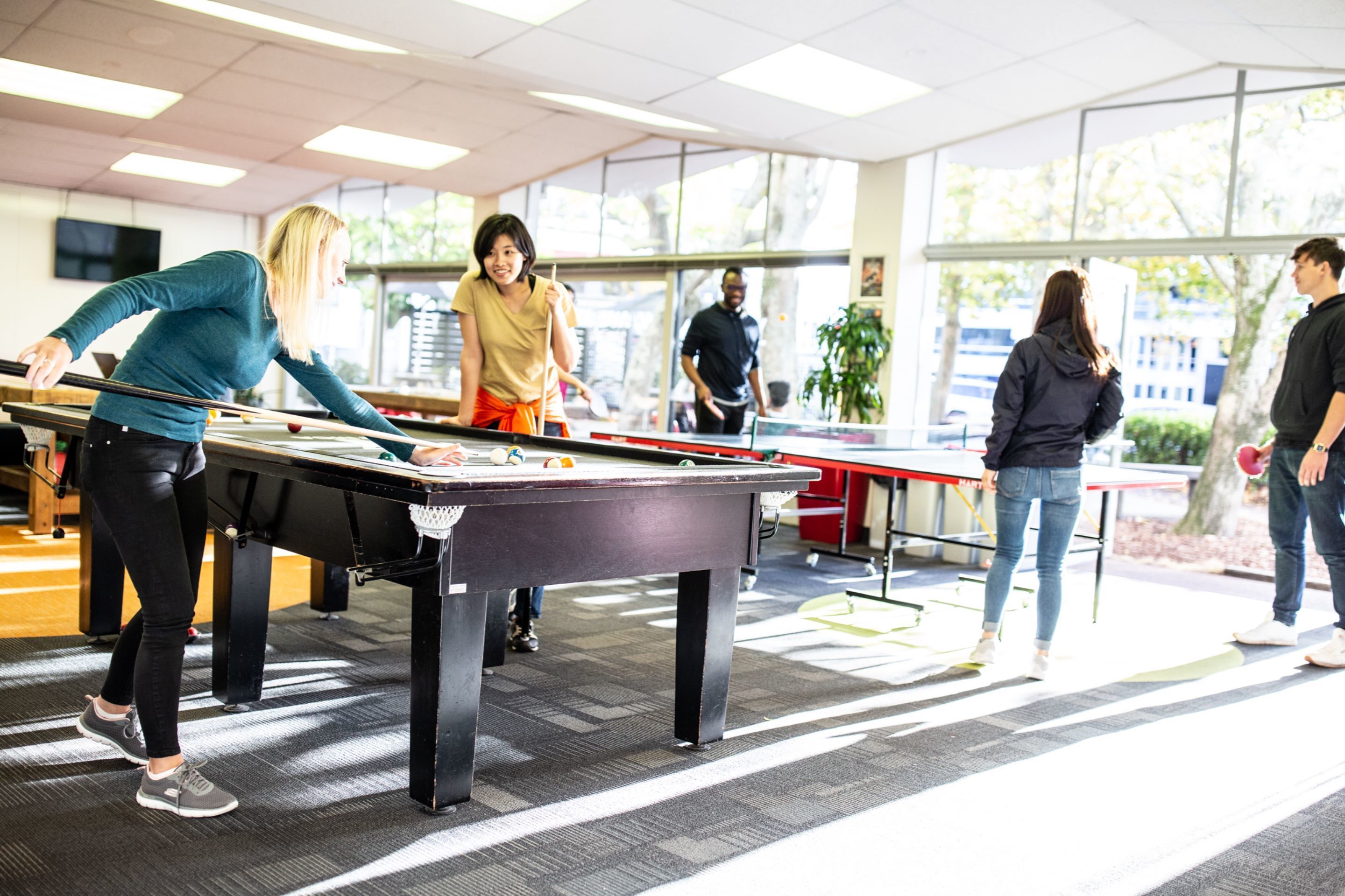 Une table de billard se trouve dans un espace intérieur bien éclairé, doté de grandes fenêtres, d’un téléviseur à écran plat fixé au mur et d’un coin salon décontracté avec des fauteuils.
