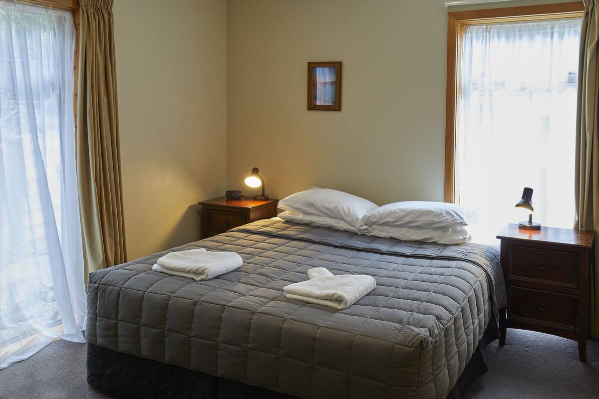 Una cama bien hecha con una colcha acolchada, dos almohadas y dos toallas dobladas. Hay una lámpara de noche sobre una mesita de madera.