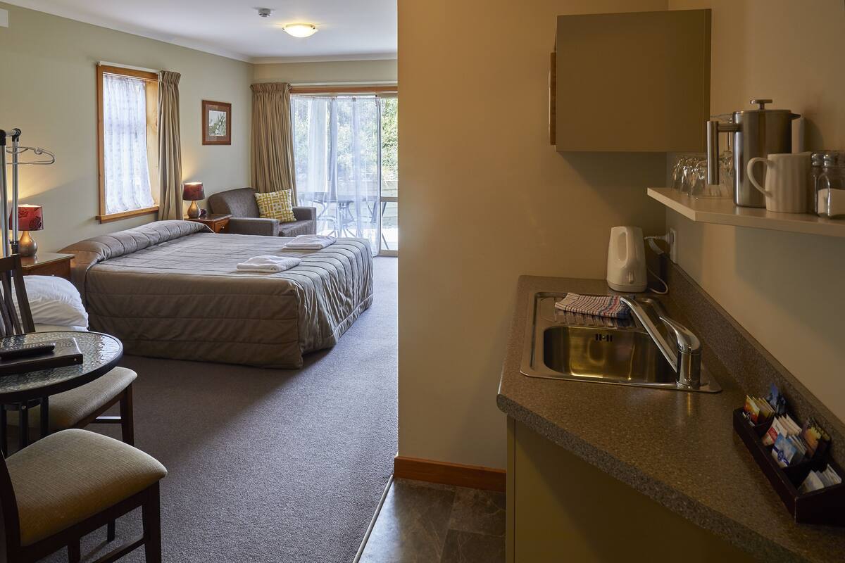 Habitación de hotel con cama, escritorio con silla, una mesita y una kitchenette con lavabo y encimera.