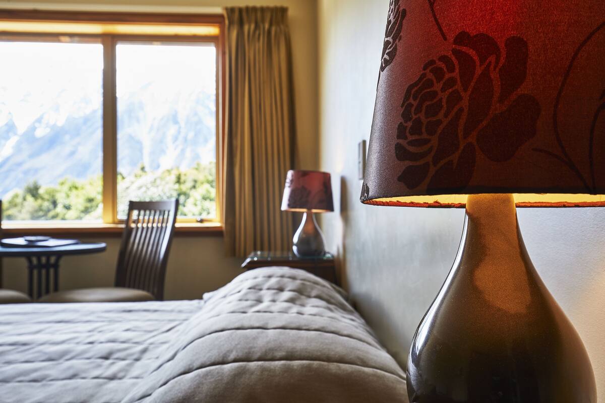 Una cama con cabecera, una lámpara de noche, una silla, una mesita y una ventana con vistas a las montañas.