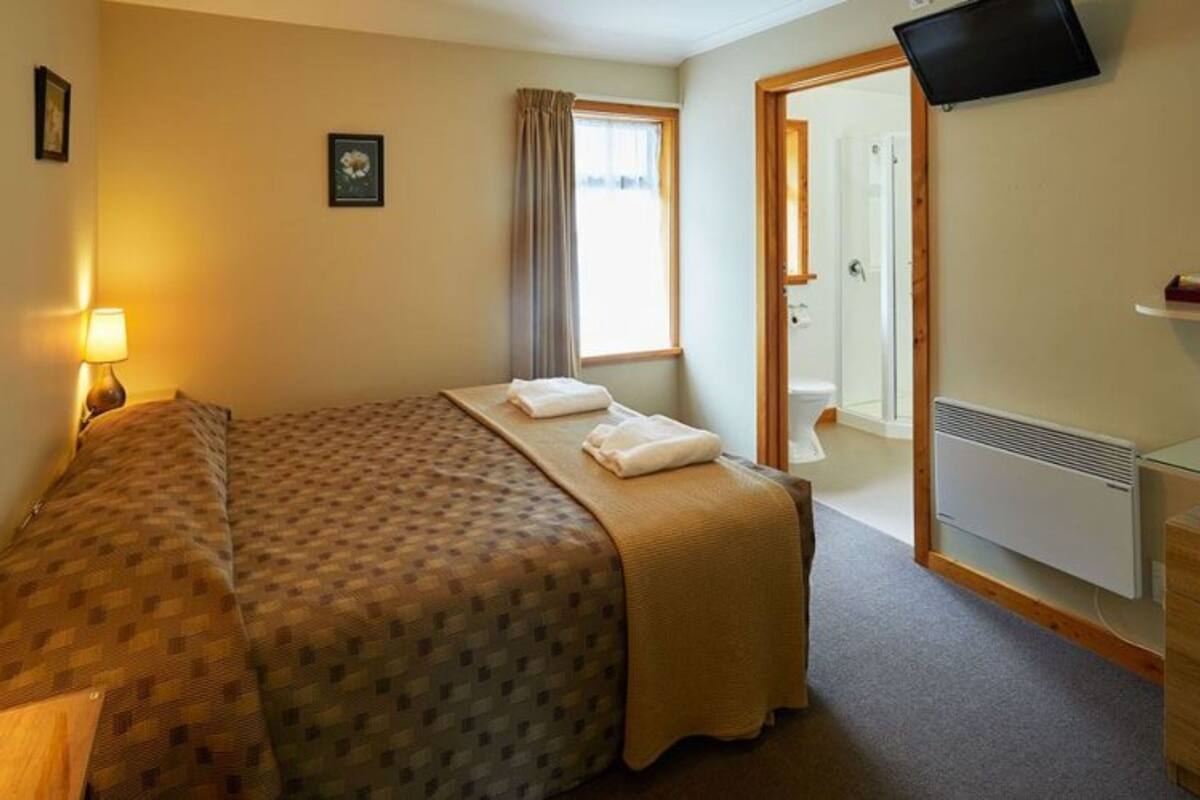 Habitación de hotel con cama, lámpara de noche, televisor de pantalla plana, ventana con cortinas y baño visible a través de una puerta abierta.