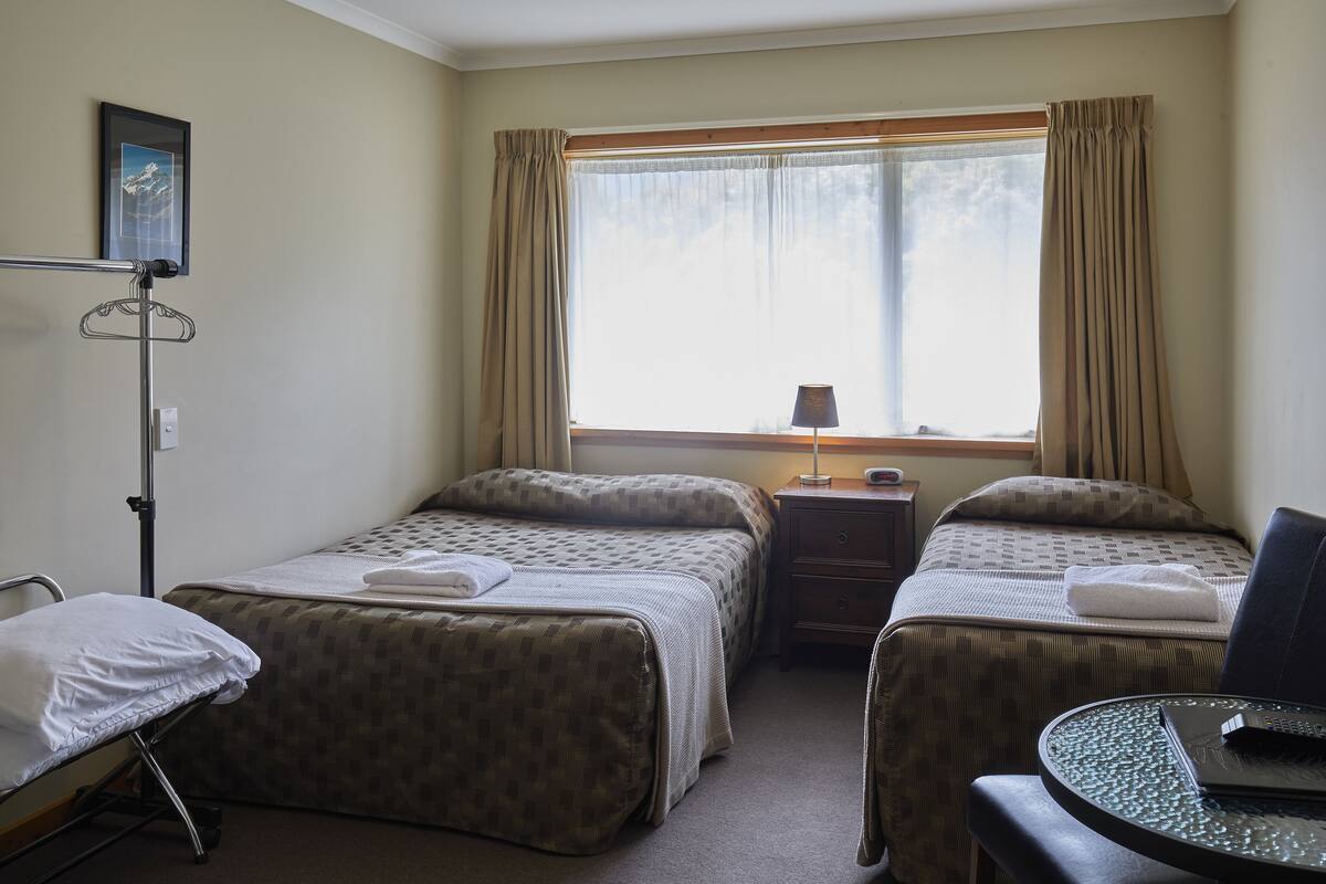 Habitación de hotel con dos camas, una mesita de noche con lámpara, una pequeña mesa con una computadora portátil y una ventana con cortinas.