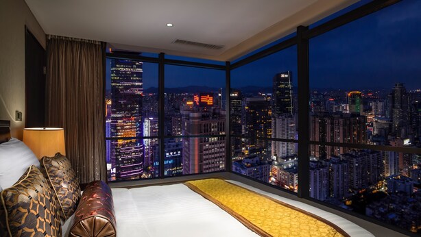 Een hotelkamer met een groot bed, uitzicht op een verlichte stad en een raam met gordijnen.