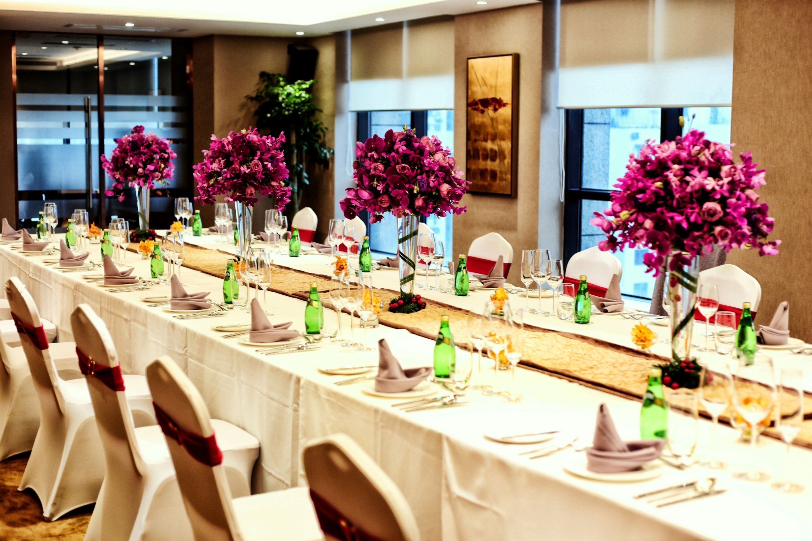 Una larga mesa de banquete vestida con manteles blancos, botellas verdes y flores púrpuras.