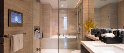 A modern bathroom with a glass-enclosed shower, a bathtub, and a toilet. There is a flat-screen TV mounted on the wall and a vase with yellow flowers on the vanity.