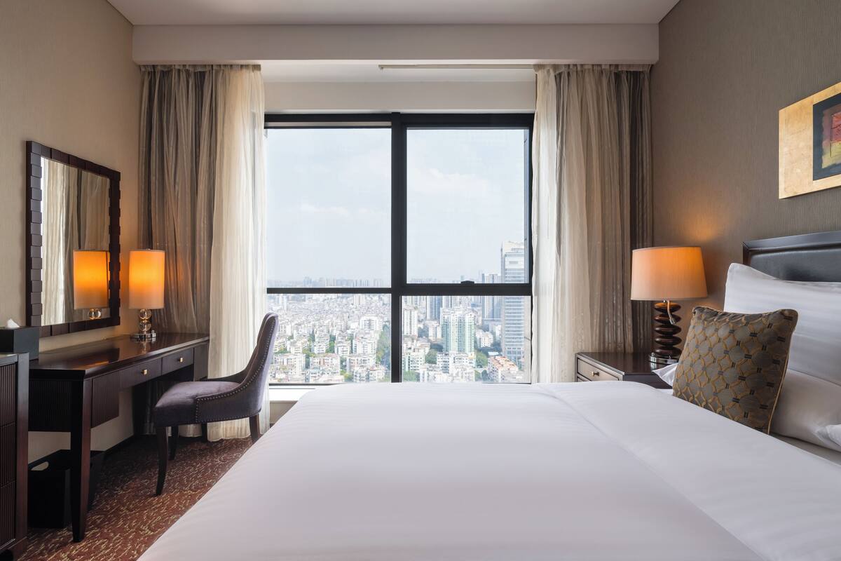 A hotel room with a large bed, a desk, a chair, and a view of a cityscape through a large window.