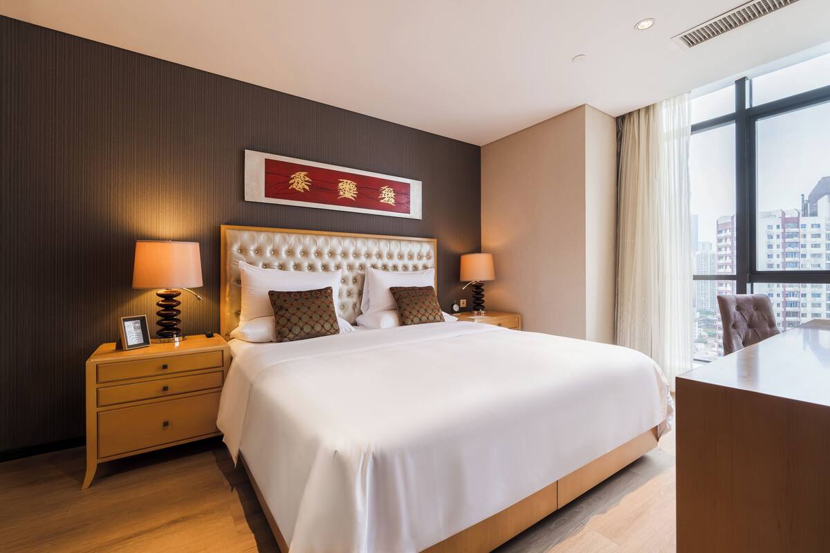 A modern hotel room with a large bed, bedside tables with lamps, a desk, and a view of the city through large windows.