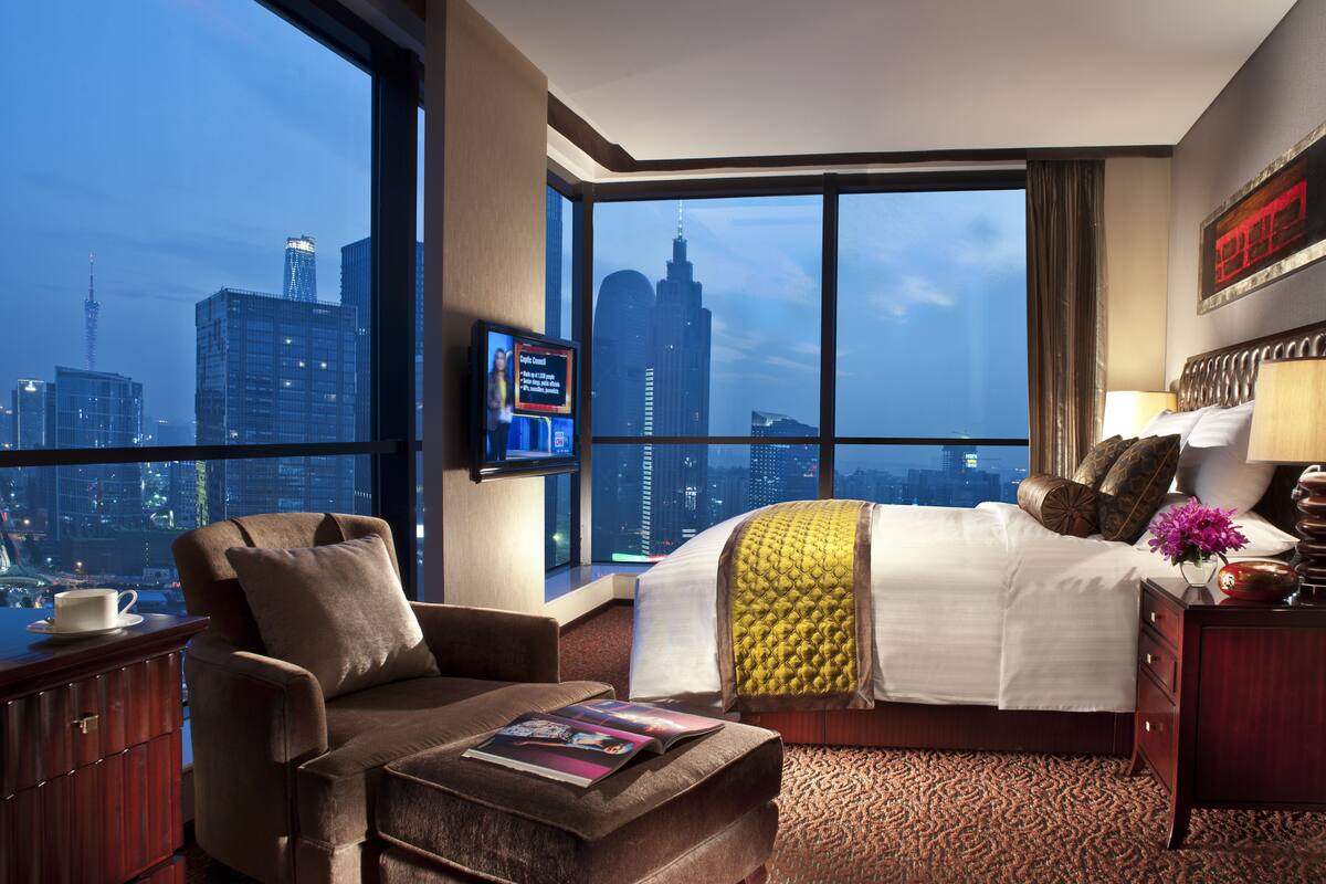 A hotel room with a large window offering a city view, a bed with a yellow accent pillow, a brown armchair, a bedside table with a lamp, and a flat-screen TV mounted on the wall.